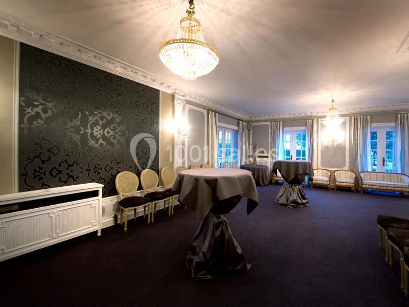 Salle élégante avec lustres, tables hautes couvertes de nappes grises, chaises et grandes fenêtres laissant entrer la…