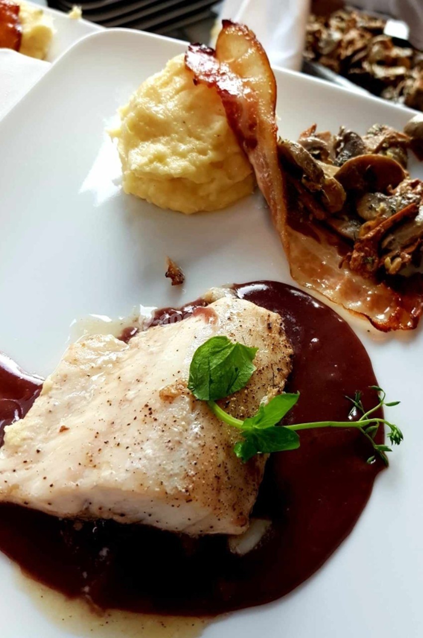 Filet de poisson avec sauce au vin rouge, purée, lard croustillant et garniture de champignons sur une assiette blanche.