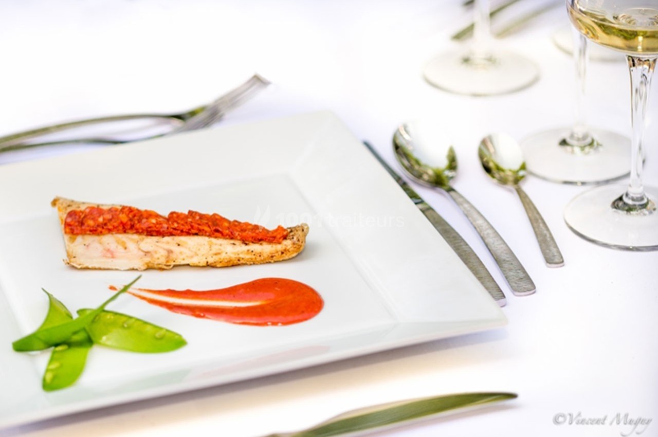 Assiette gastronomique avec tranche de poisson, purée rouge, pois gourmands et couverts sur une table blanche.