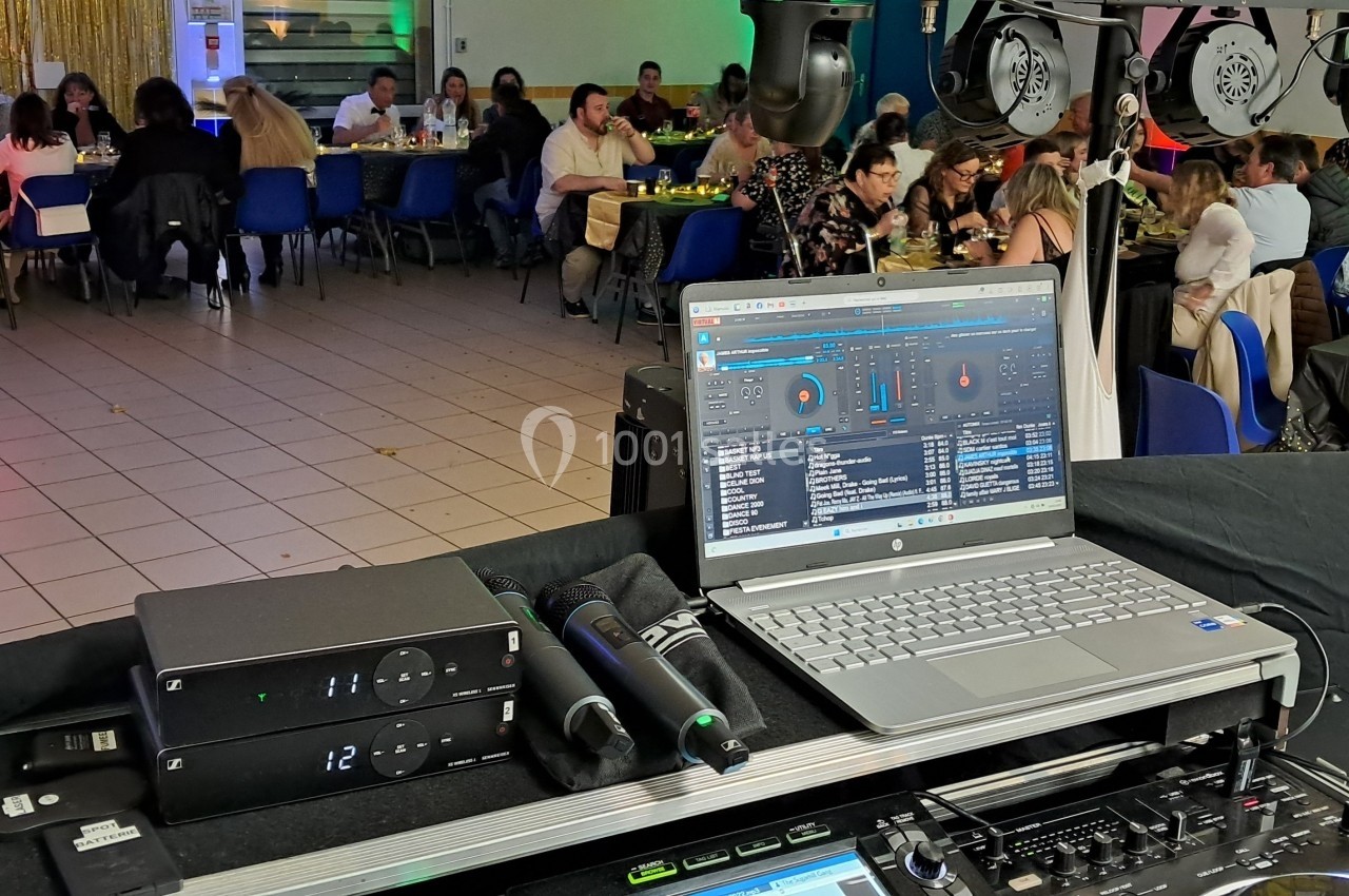 Vue d'une salle de réception avec des invités attablés et un poste de DJ en premier plan, équipé d'un ordinateur portable.