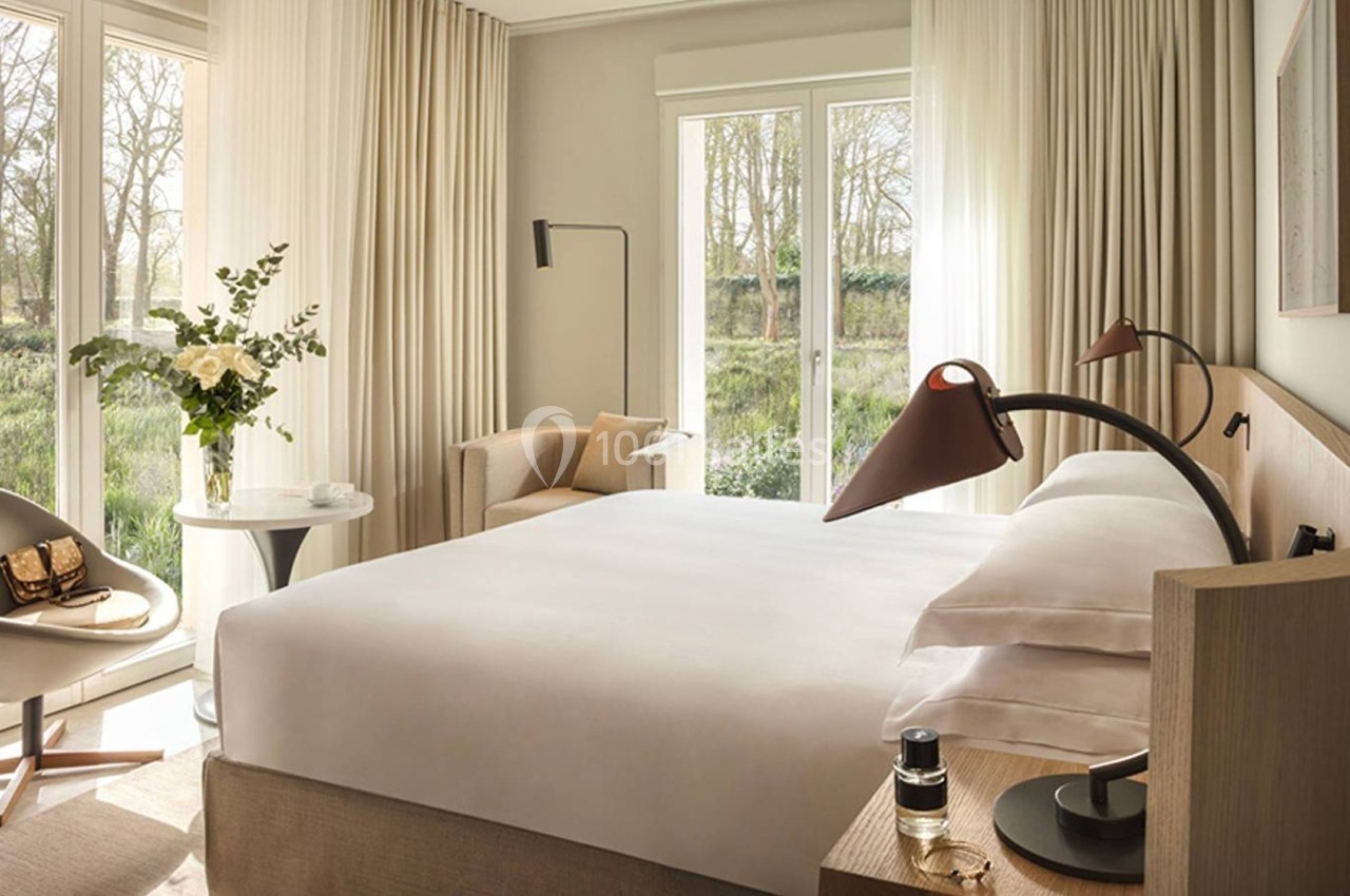 Chambre lumineuse avec lit double, fauteuil, table avec vase de fleurs, lampes modernes et vue sur un jardin verdoyant.