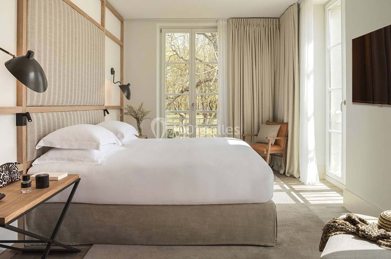 Chambre lumineuse avec lit double, bureau en bois, fauteuil, grande fenêtre et rideaux beiges laissant entrer la lumière.