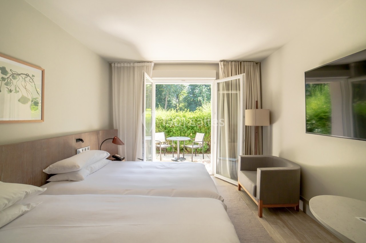 Chambre lumineuse avec deux lits, un fauteuil, une télévision et une porte-fenêtre donnant sur une terrasse et un jardin.