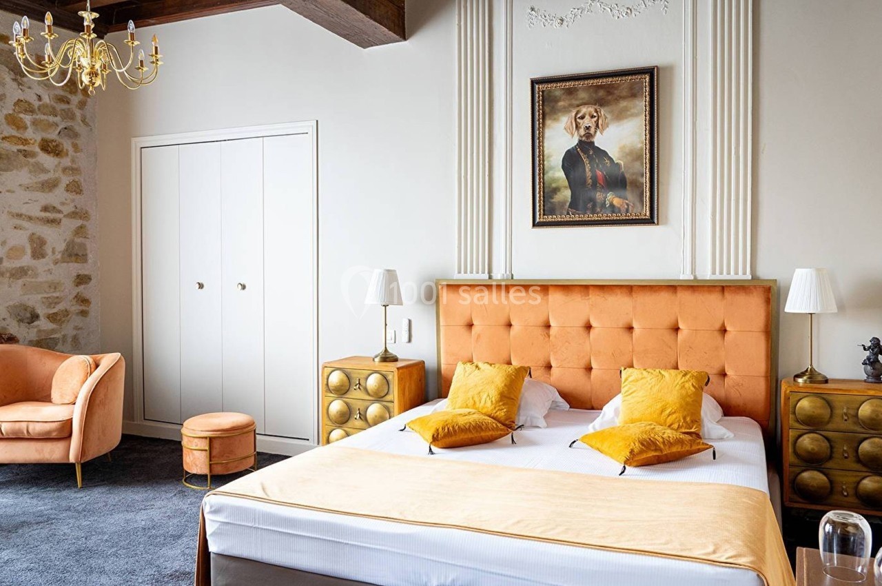 Chambre lumineuse avec lit double, tête de lit orange, coussins jaunes, mobilier rétro et portrait au mur.