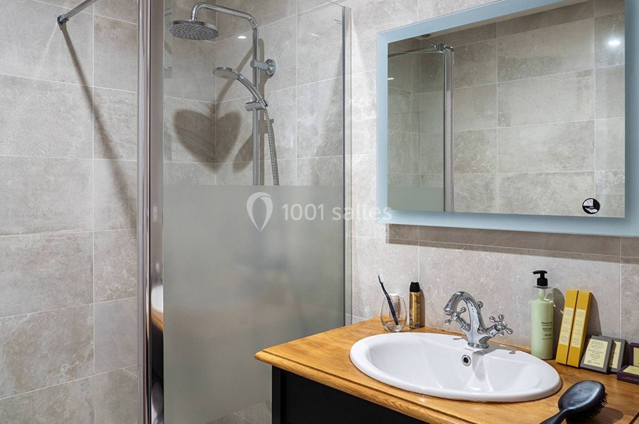 Salle de bain avec douche à paroi vitrée, lavabo sur meuble en bois, miroir éclairé et accessoires de toilette.