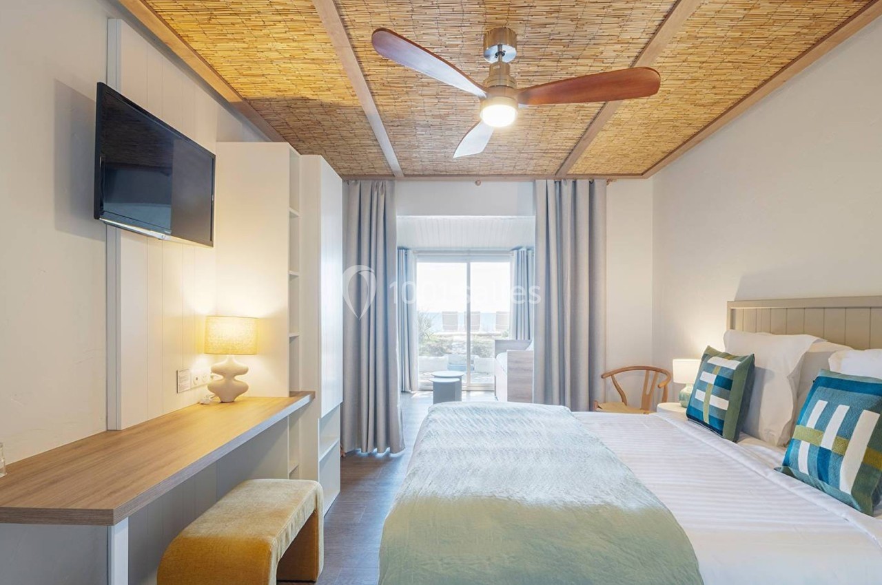 Chambre lumineuse avec lit double, coussins colorés, bureau, télé murale, ventilateur au plafond et balcon avec vue.