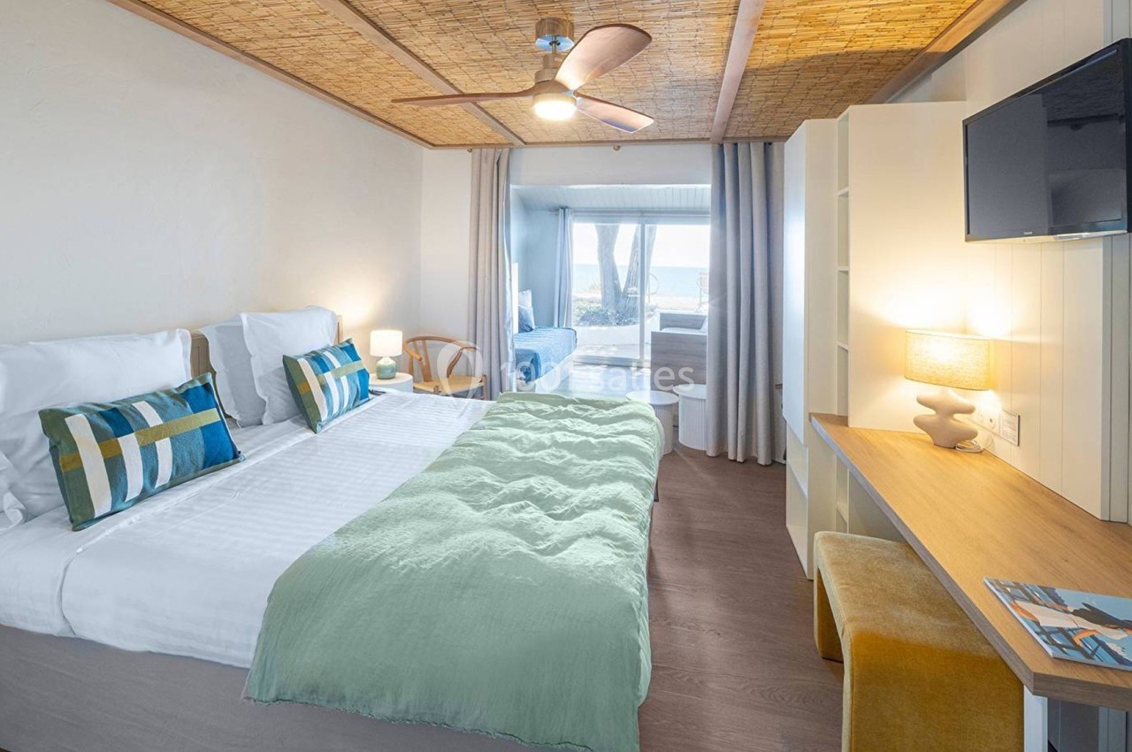 Chambre lumineuse avec lit double, bureau, télévision murale, plafond en bois et vue sur l'extérieur à travers une baie…