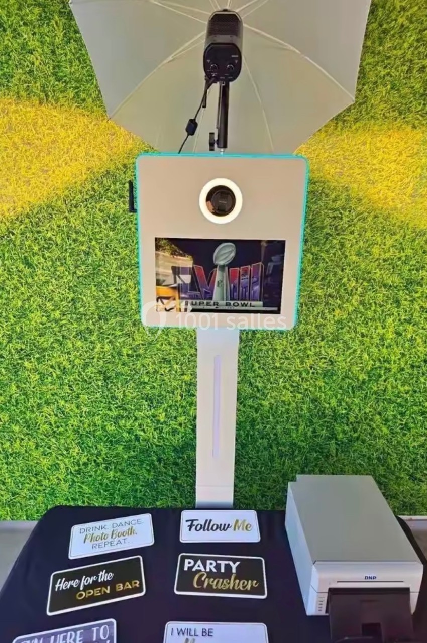 Borne photo avec éclairage, écran et accessoires humoristiques posés devant un mur végétal.