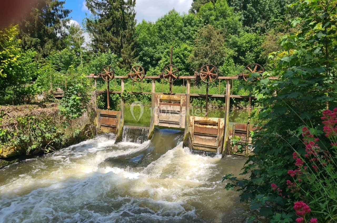 Écluse en bois avec roues dentées, entourée de végétation dense, laissant passer de l'eau en mouvement rapide.