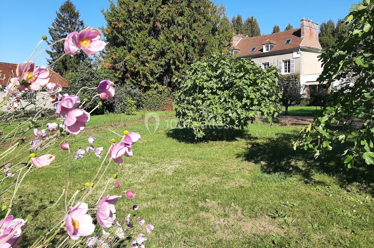 Jardin verdoyant avec des fleurs roses au premier plan, des arbres et une maison en pierre sous un ciel bleu.