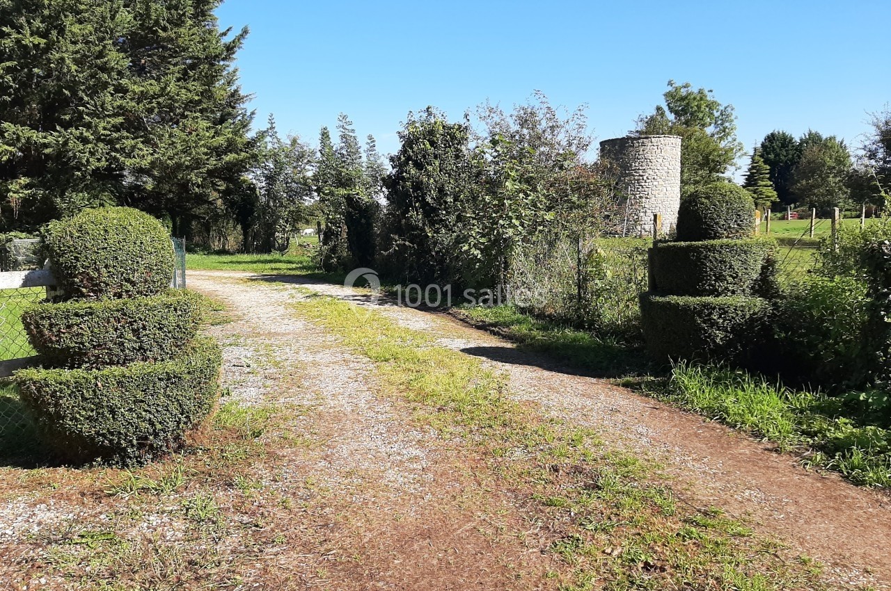Chemin de gravier bordé de buissons taillés, menant à une tour en pierre dans un paysage verdoyant.