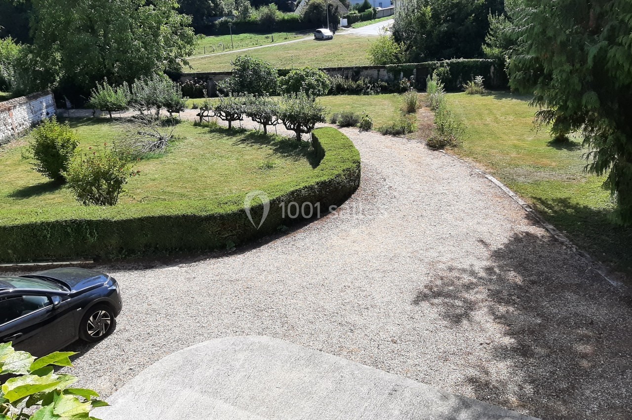 Allée en gravier bordée de pelouses et d'arbustes, avec une voiture garée à gauche et un paysage verdoyant en arrière-plan.