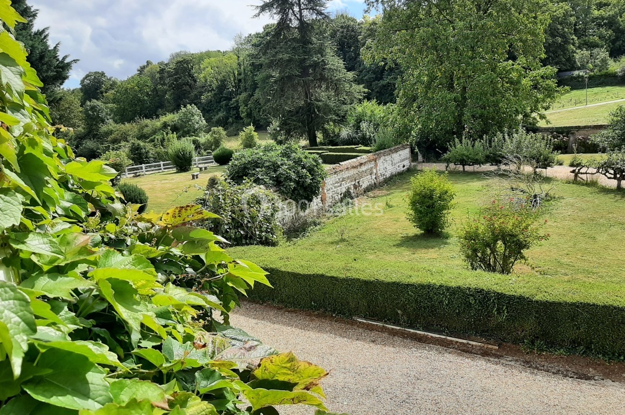 Jardin verdoyant avec pelouse, arbres et haies, bordé par un chemin de gravier et un mur en pierre.