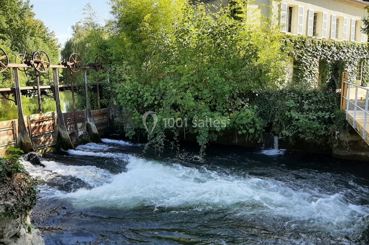Vue d'un cours d'eau avec un petit barrage, entouré de végétation et d'une maison en arrière-plan.