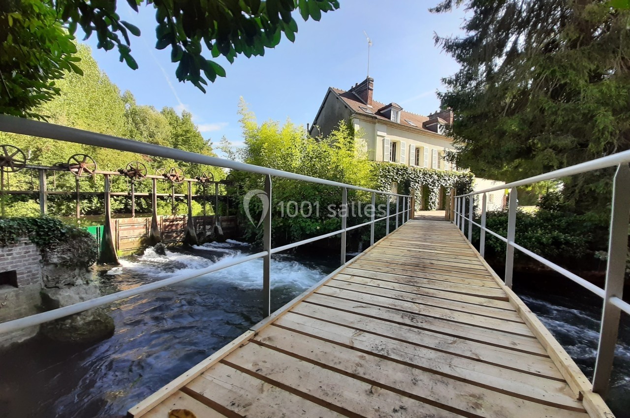 Passerelle en bois traversant un cours d'eau près d'une maison entourée de végétation dense.