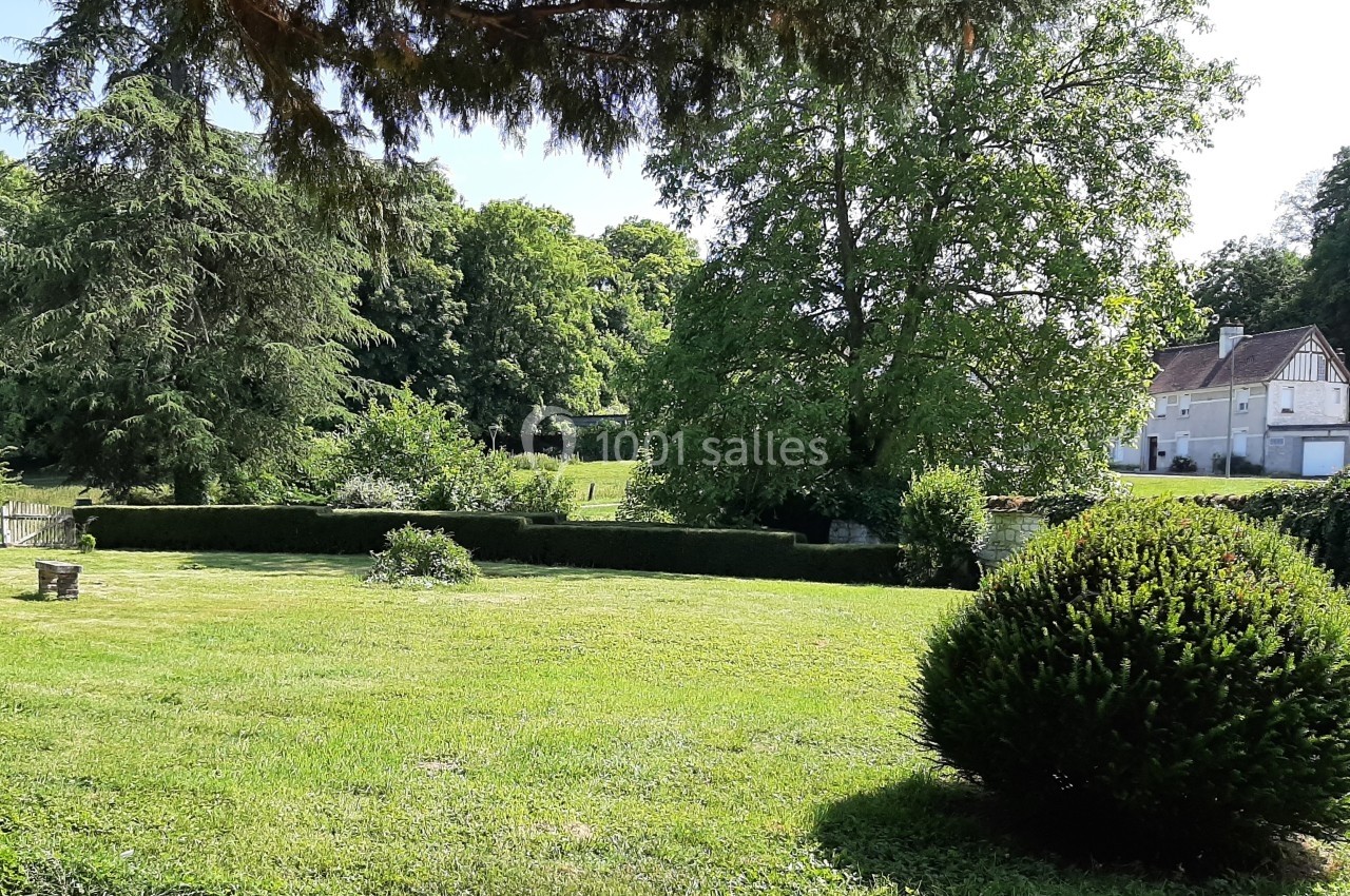 Jardin verdoyant avec pelouse, arbustes et grands arbres, maison en pierre visible en arrière-plan.