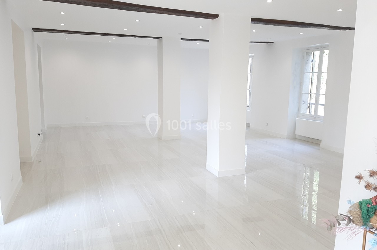 Salle lumineuse avec sol en carrelage clair, murs blancs, poutres apparentes et grandes fenêtres laissant entrer la lumière…