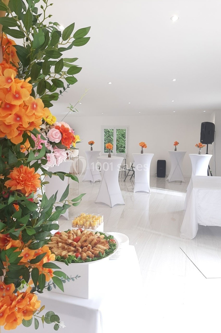 Salle lumineuse décorée de fleurs orange et roses, avec buffet et tables hautes habillées de nappes blanches.