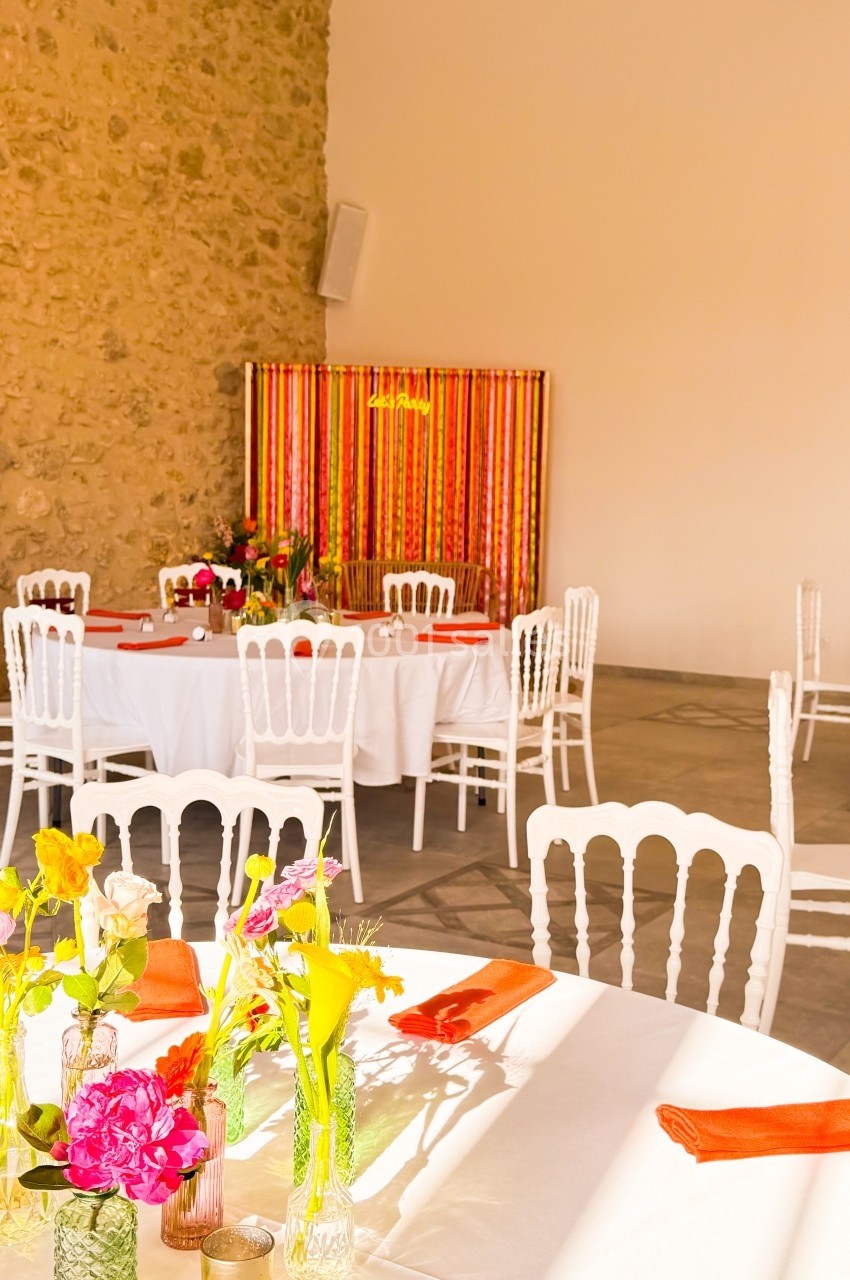 Salle décorée pour un événement avec tables rondes, nappes blanches, chaises blanches et fleurs colorées en centre de table.
