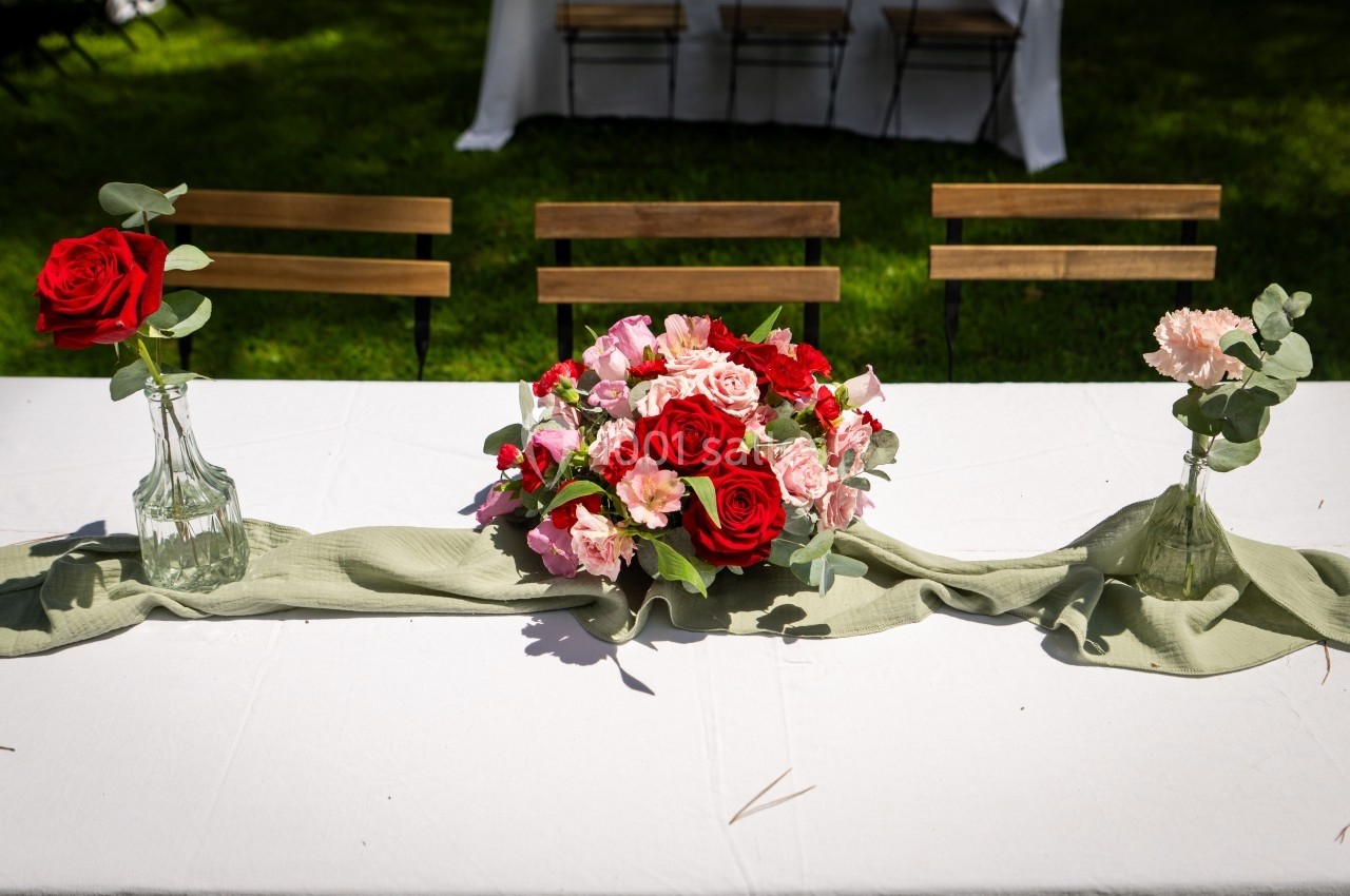 Centre de table avec bouquet de roses rouges et roses, entouré de vases en verre sur une nappe blanche en extérieur.