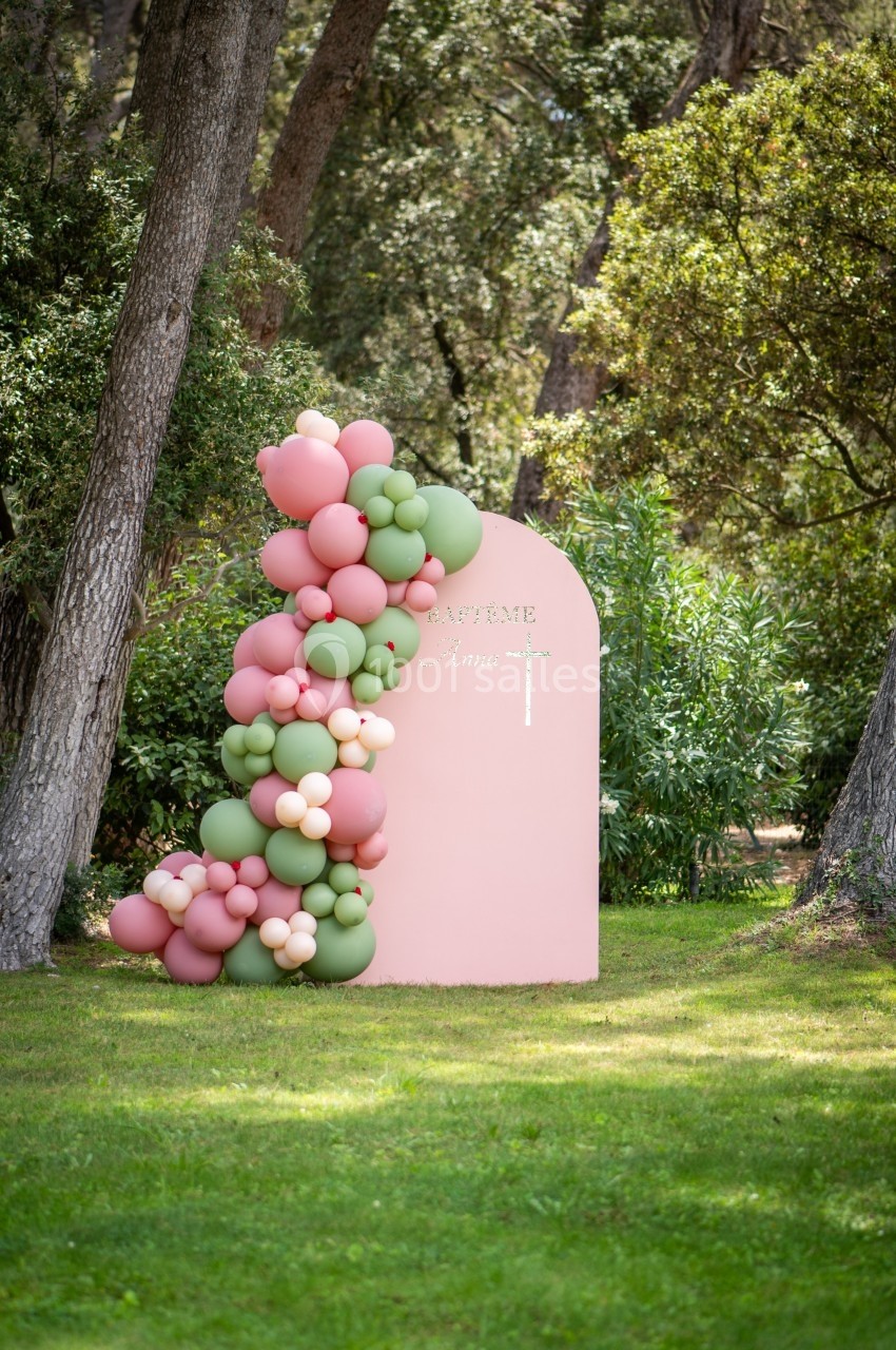 Décoration extérieure avec arche rose ornée de ballons verts et roses, entourée d'arbres et de végétation.