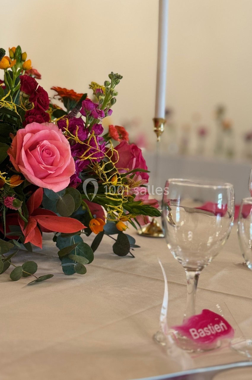 Centre de table floral coloré avec des roses et des verres disposés sur une nappe blanche, bougie en arrière-plan.