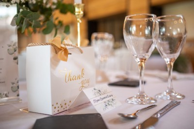 Salle de réception décorée pour un mariage, avec tables dressées, chandeliers suspendus et poutres apparentes au plafond.