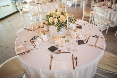 Salle de réception décorée pour un mariage, avec tables dressées, chandeliers suspendus et poutres apparentes au plafond.