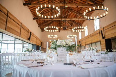 Salle de réception décorée pour un mariage, avec tables dressées, chandeliers suspendus et poutres apparentes au plafond.