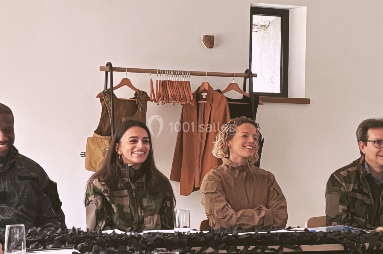 Des personnes en tenue militaire assises autour d'une table, souriantes, avec des vêtements suspendus en arrière-plan.