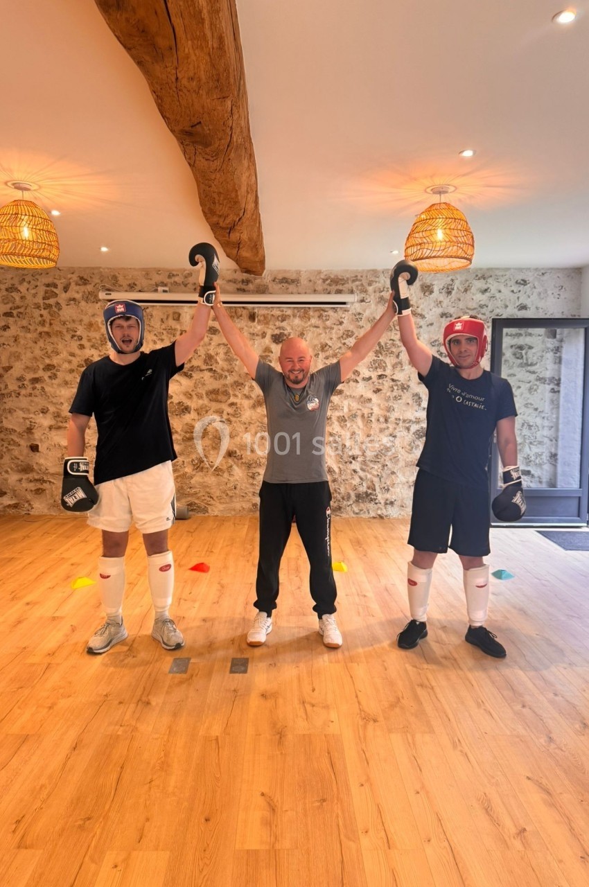 Deux personnes en équipement de boxe posent avec un entraîneur levant leurs bras dans une salle au sol en bois.