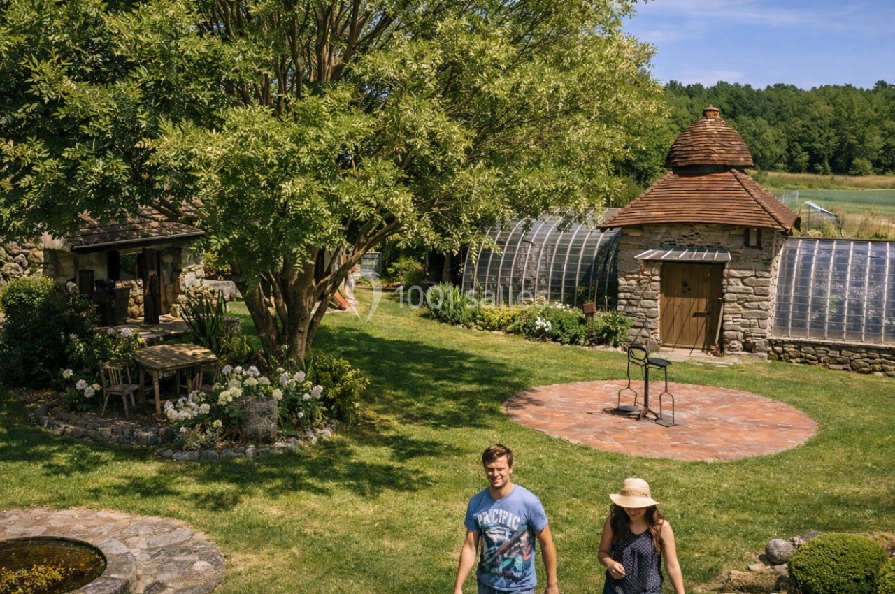 Un jardin verdoyant avec des serres, une petite maison en pierre et deux personnes marchant sur une pelouse.