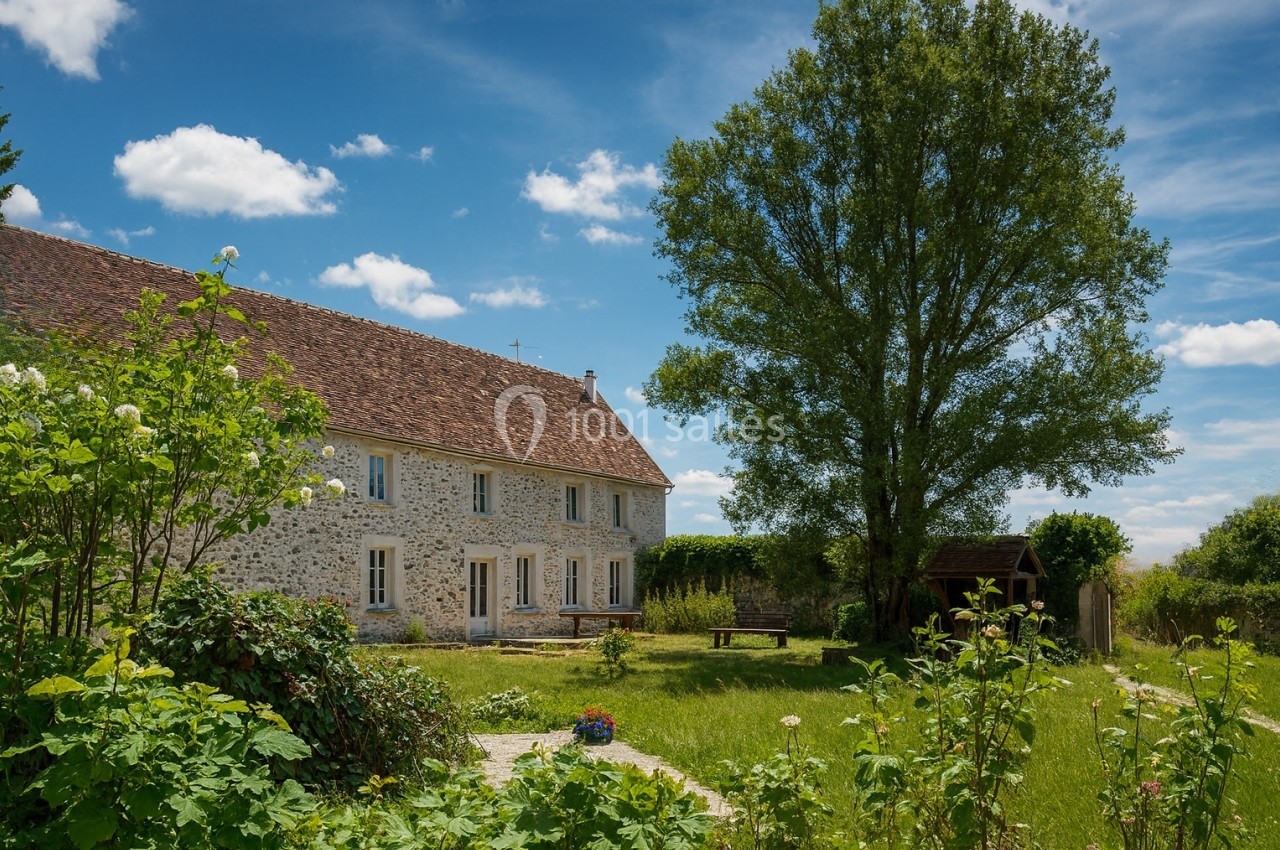Maison en pierre avec toit en tuiles, entourée d'un jardin verdoyant et d'arbres sous un ciel bleu.
