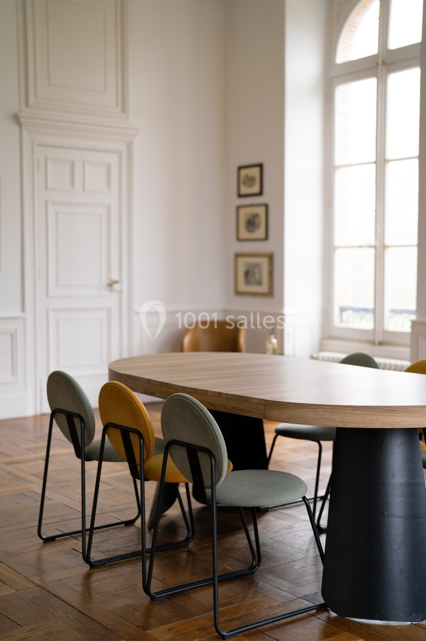 Salle lumineuse avec une grande table en bois entourée de chaises modernes, parquet au sol et fenêtres hautes.