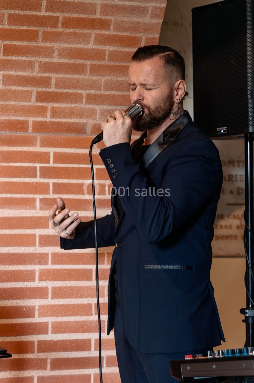 Un homme en costume sombre chante au micro devant un mur en briques, avec un haut-parleur en arrière-plan.