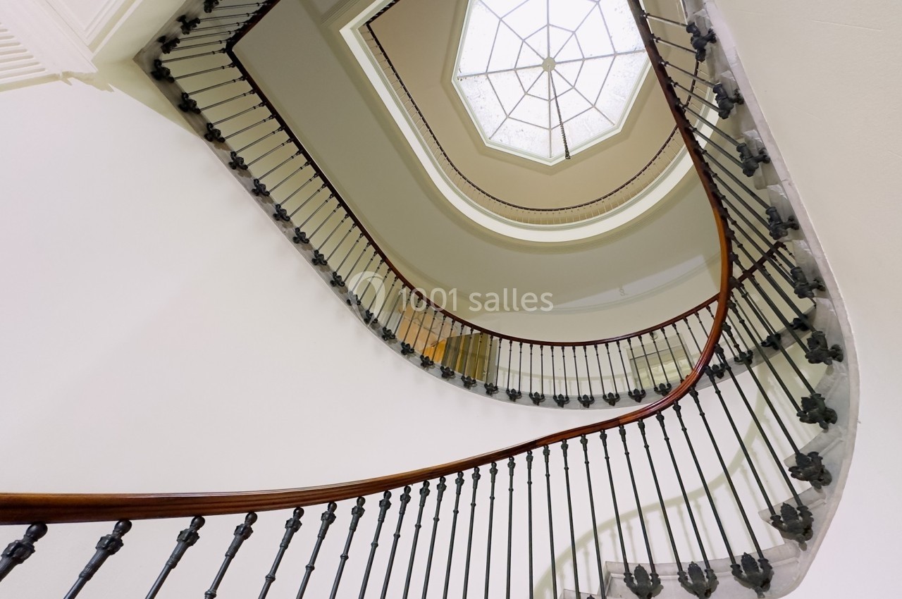 Escalier en colimaçon vu d'en bas, avec une rampe en bois et une verrière hexagonale au plafond.