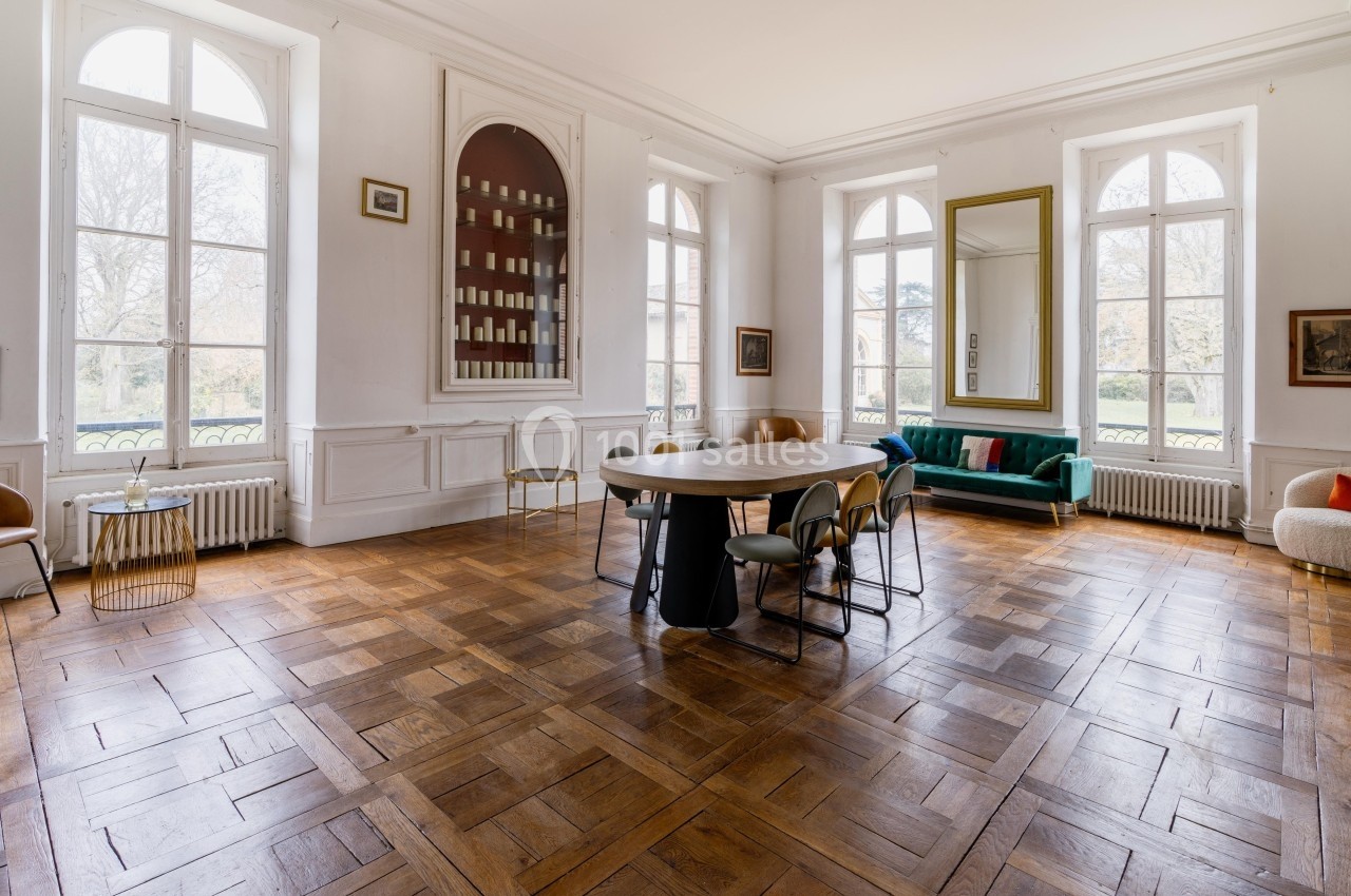 Salle lumineuse avec parquet en bois, table ovale entourée de chaises, canapé vert et grandes fenêtres donnant sur un jardin.