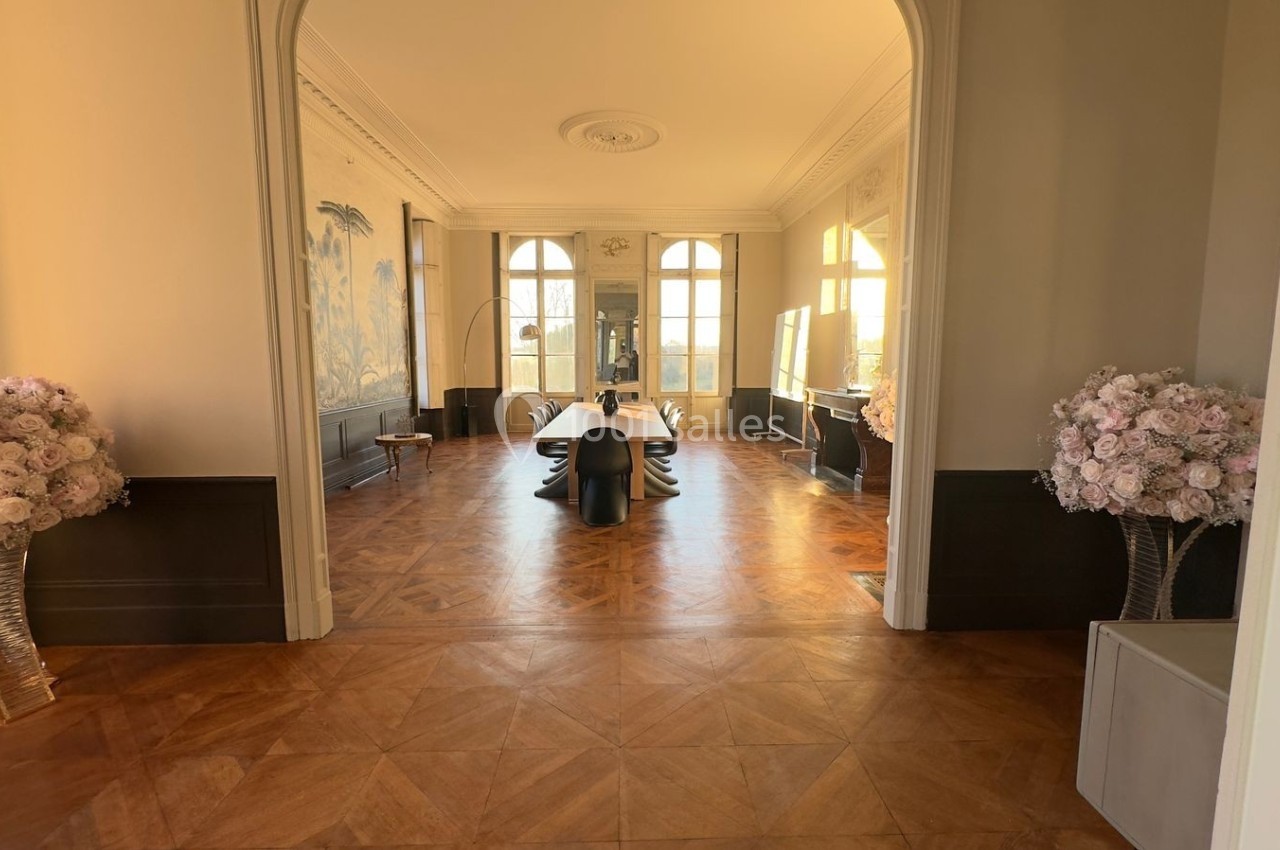 Salle lumineuse avec parquet en bois, grande table noire centrale, murs décorés et bouquets de fleurs roses.