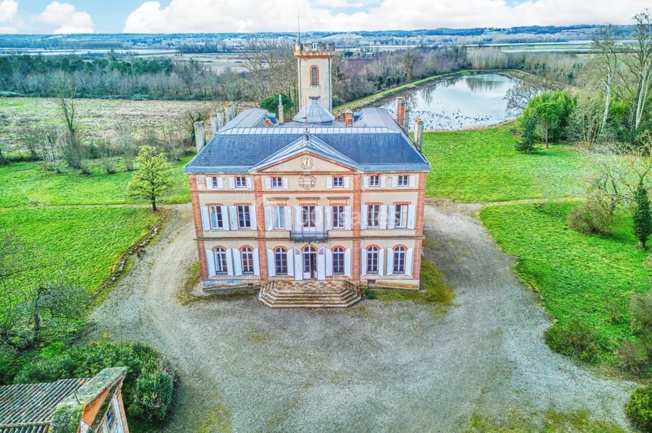 Vue aérienne d'un château en briques entouré de verdure, avec un étang en arrière-plan.