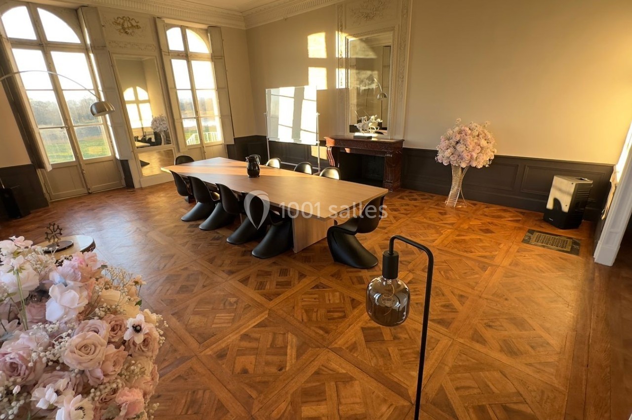 Salle lumineuse avec parquet en bois, grande table entourée de chaises noires, décorée de fleurs et baignée de lumière…