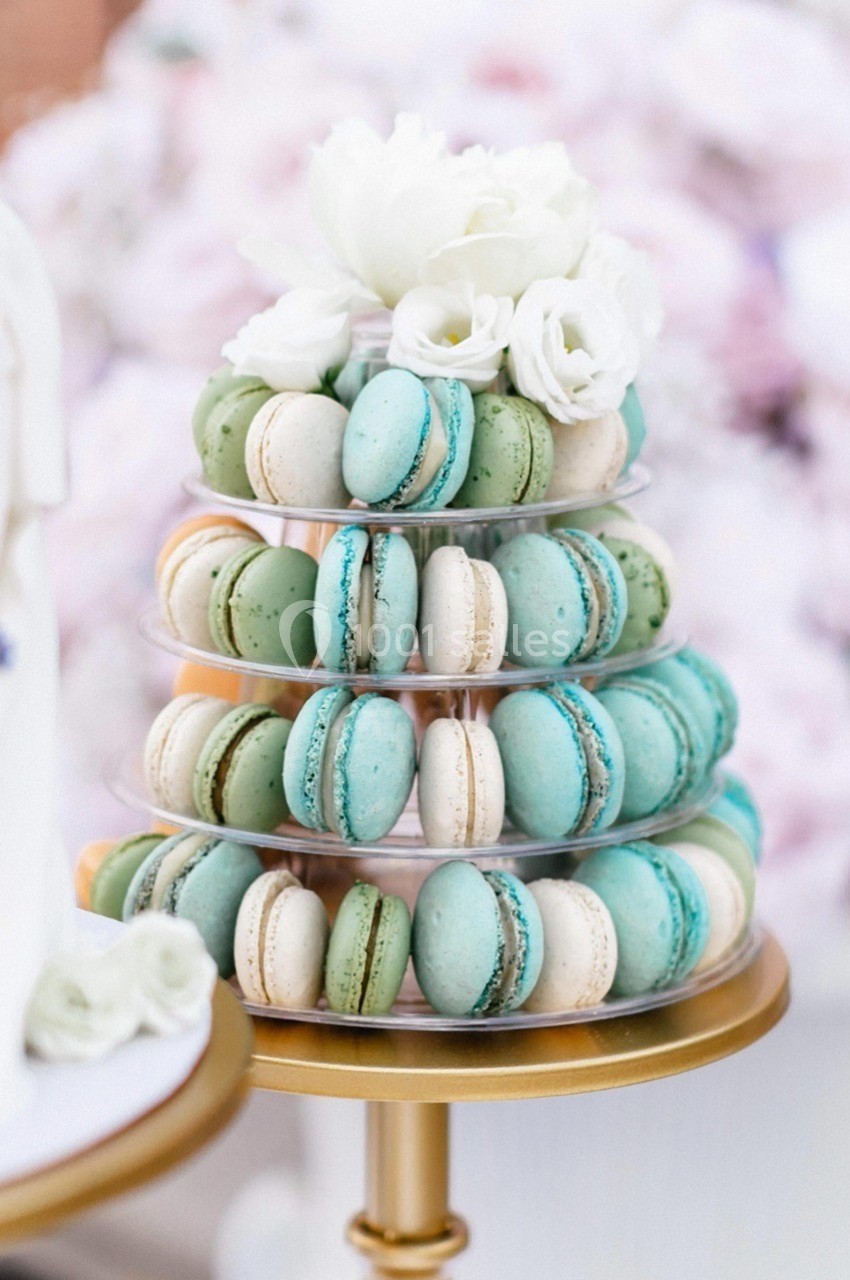 Tour de macarons pastel verts et bleus sur un présentoir doré, décorée de fleurs blanches en haut.