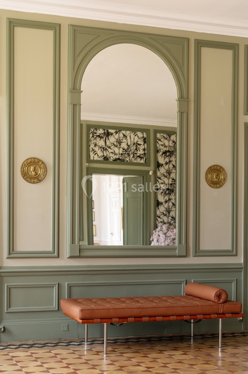 Miroir encadré dans une pièce aux murs vert olive, avec une banquette en cuir brun et un sol en carreaux anciens.