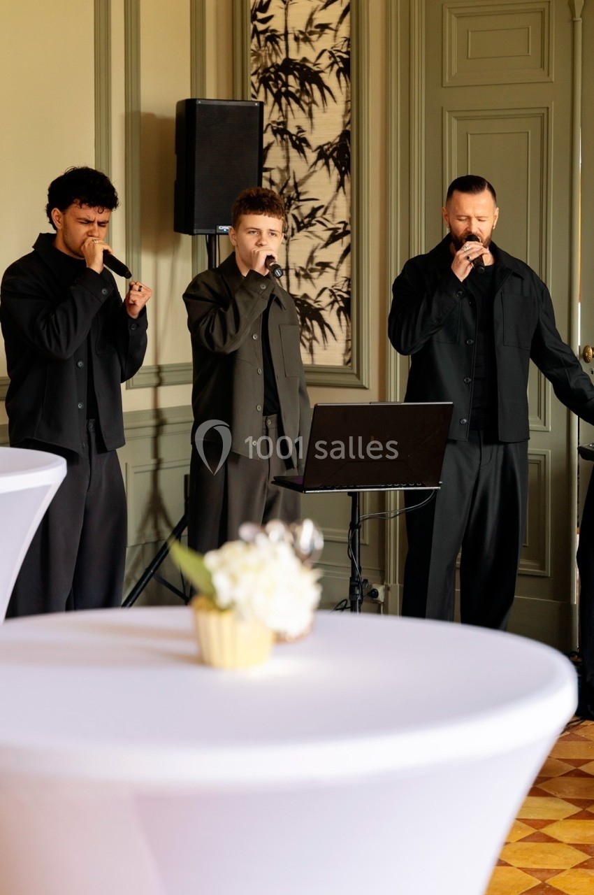 Trois hommes chantant avec des micros dans une salle élégante, devant des tables décorées de fleurs.
