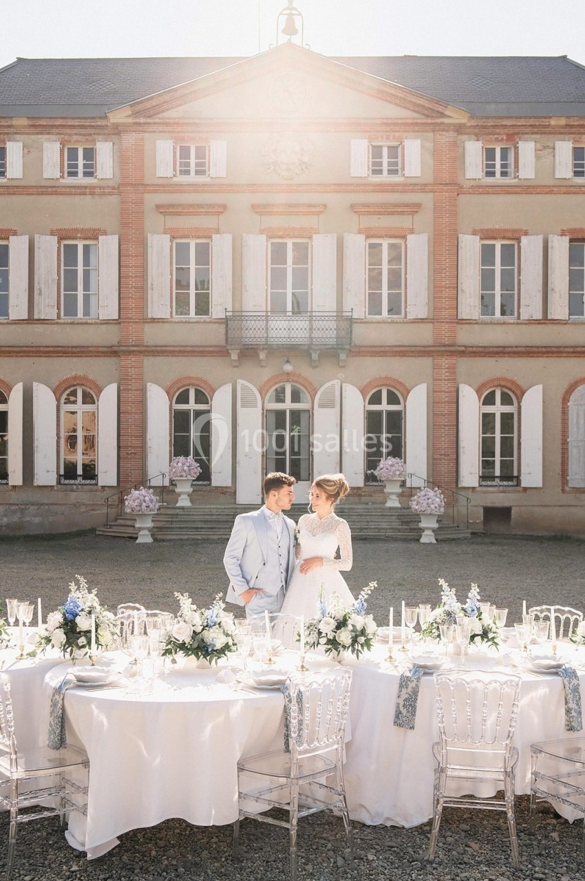 Un couple debout devant une table de réception élégamment dressée, avec un manoir en arrière-plan.