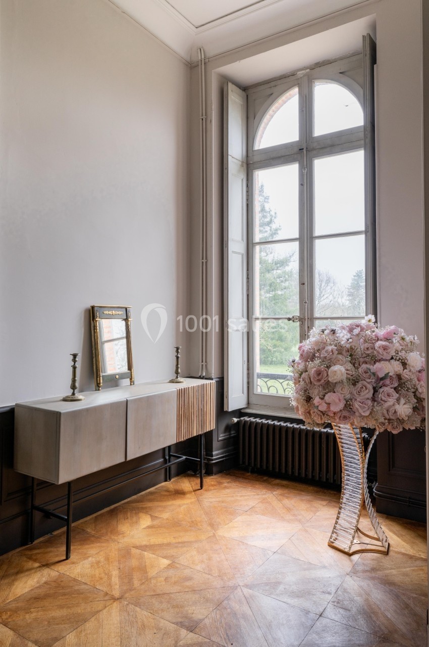 Console moderne avec bougeoirs et miroir, bouquet de fleurs sur pied en verre, près d'une fenêtre à battants.