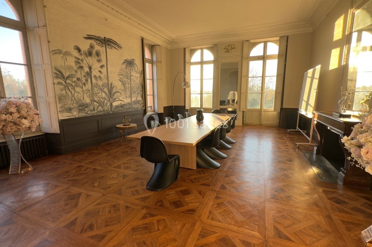 Salle de réunion lumineuse avec grande table en bois, chaises noires, parquet en chevrons et décor mural tropical.