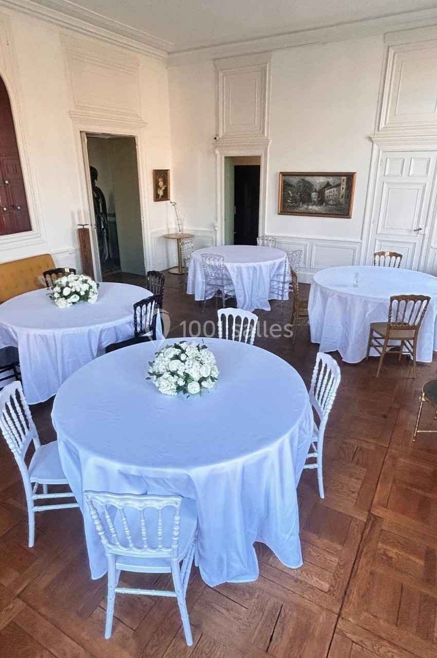 Salle avec parquet, tables rondes couvertes de nappes blanches, chaises variées et bouquets de fleurs blanches.