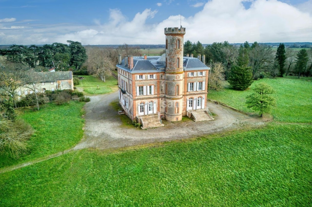 Château en briques rouges avec une tour centrale, entouré de pelouses et d'arbres sous un ciel partiellement nuageux.