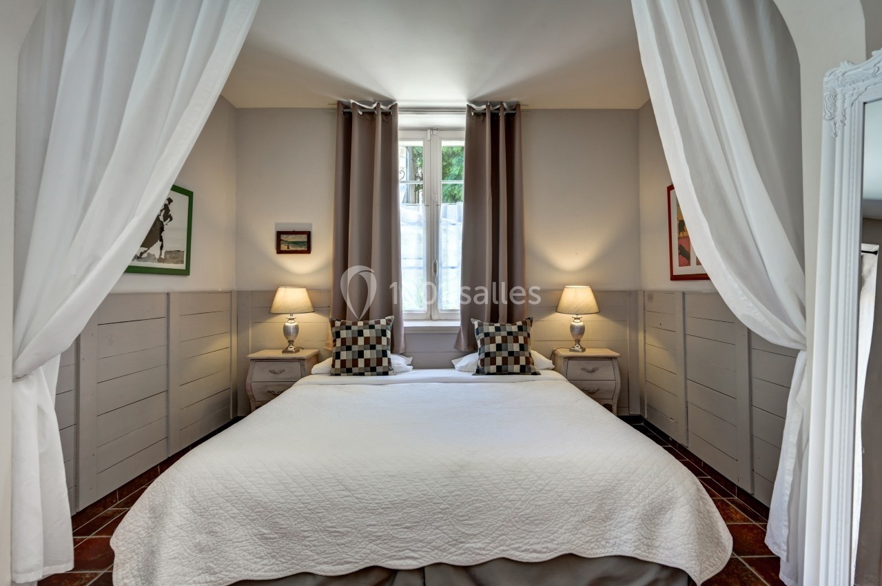 Lit double avec couvre-lit blanc, encadré par deux tables de chevet et lampes, dans une chambre lumineuse avec rideaux…