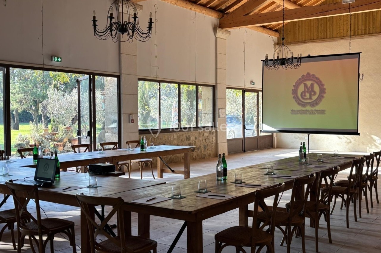Salle de réunion lumineuse avec grandes tables en bois, chaises, écran de projection et vue sur un extérieur arboré.